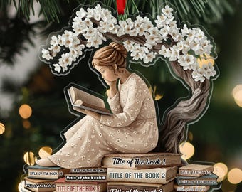 May include: Clear acrylic ornament featuring a woman reading under a flowering tree. The woman is seated on a stack of books with titles like "Title of the book 1" and "Title of the book 2". A red ribbon is attached for hanging.