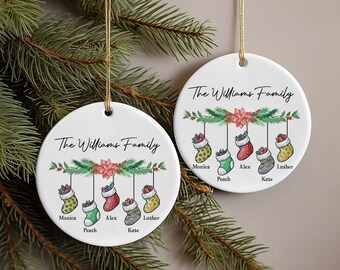 May include: Two round white ceramic Christmas ornaments with the text "The Williams Family" and a green garland with red poinsettia and five stockings with names written below each stocking.