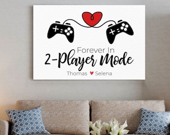 May include: A white canvas print featuring two black video game controllers connected by a red heart. The text reads "Forever In 2-Player Mode" with the names "Thomas" and "Selena" below. The print is displayed above a sofa.