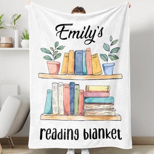 May include: White fleece blanket with the text "Emily's reading blanket" printed in black. The blanket features a watercolor illustration of two shelves with books and potted plants.