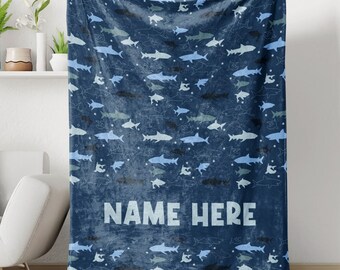 May include: A blue fleece blanket with a white outline of sharks and stars on a dark blue background. The blanket has the text "NAME HERE" in white.