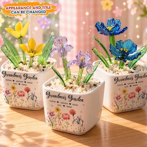 May include: Three white square ceramic pots with glass flowers. Each pot has a floral design and the text "Grandma's Garden" with names. The flowers are yellow, purple, and blue. The pots are filled with small white stones.