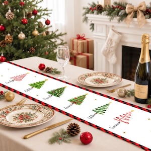 May include: A white table runner featuring a red and black buffalo plaid border and a repeating pattern of six illustrated Christmas trees. Each tree is personalized with a name: Emily, Adela, David, Kenna, Chloe, and Donna, accented by gold stars.