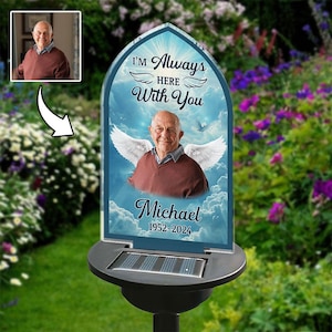 May include: A solar-powered memorial garden stake with a photo of a man, angel wings, and the text "I'm Always Here With You" and "Michael 1952-2024". The stake is set in a garden with flowers.