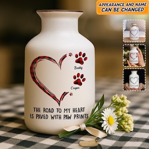 May include: White ceramic vase with a brown rim, featuring a heart design in red plaid and paw prints. The text reads "THE ROAD TO MY HEART IS PAVED WITH PAW PRINTS." Includes the names "Buddy" and "Cooper."