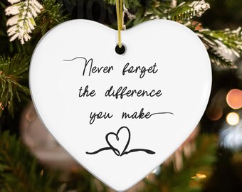May include: A white heart-shaped ornament with the message "Never forget the difference you make" in black script. A gold string is attached for hanging. The ornament is displayed on a Christmas tree.