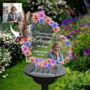 May include: A solar-powered garden stake featuring a hummingbird and floral design. The stake includes a photo and the text "Hummingbirds Appear When Angels Are Near." The base is black.