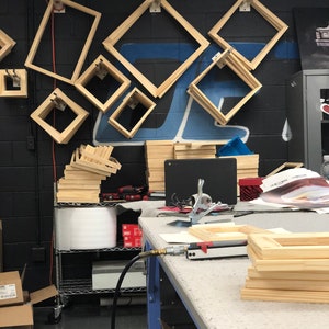 May include: A workshop table with a variety of wooden picture frames stacked on top of each other. The frames are made of natural wood and are ready to be assembled. There is a pneumatic staple gun on the table, along with other tools.