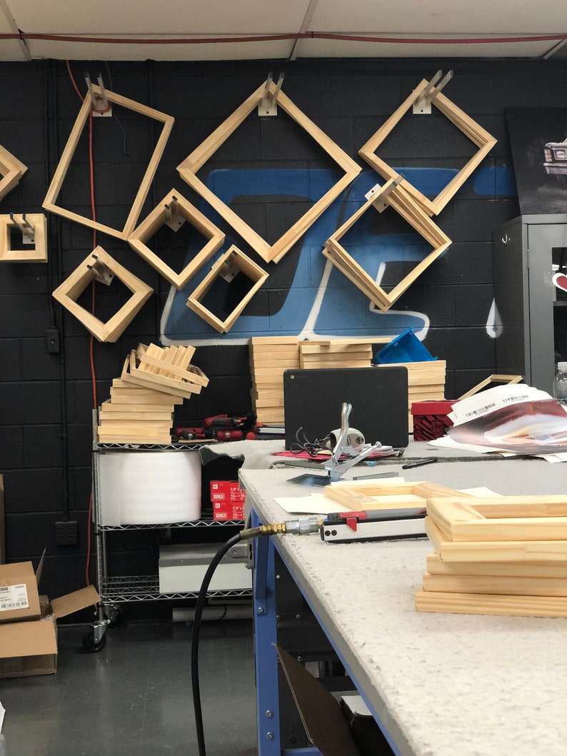 May include: A workshop table with a variety of wooden picture frames stacked on top of each other. The frames are made of natural wood and are ready to be assembled. There is a pneumatic staple gun on the table, along with other tools.