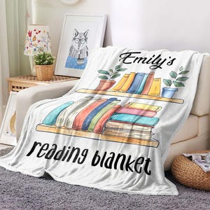 K&ouml;nnte beinhalten: Wei&szlig;e Fleece-Decke mit dem Text "Emily's reading blanket" und einer Zeichnung eines B&uuml;cherregals mit B&uuml;chern und Topfpflanzen.