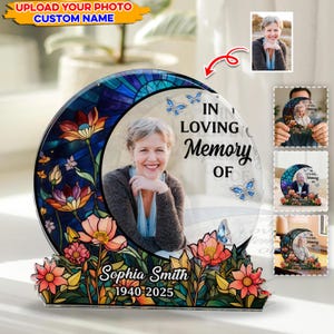 Pode incluir: Placa memorial com a foto de uma mulher, rodeada por um design de vitral de flores e uma lua crescente. O texto diz "In Loving Memory Of" e o nome "Sophia Smith 1940-2025". Borboletas também estão presentes.