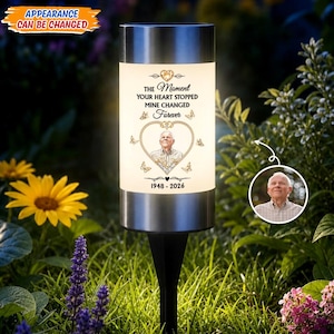 May include: A cylindrical solar garden light with a silver top and black stake. The white surface features a gold heart frame containing a portrait of an older person, surrounded by butterflies and the text: THE MOMENT YOUR HEART STOPPED MINE CHANGED FOREVER 1948-2026.
