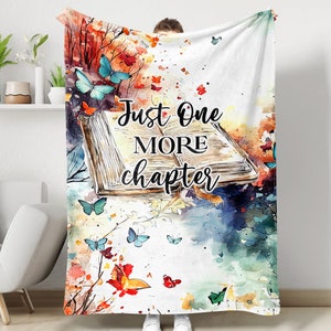 May include: A white fleece blanket with a watercolor design of an open book, butterflies, and leaves. The text "Just One More Chapter" is written in black on the book.