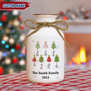 Puede incluir: Jarrón de cerámica blanca con borde marrón y lazo de yute. El jarrón presenta diseños de árboles de Navidad con nombres familiares y el texto "The Smith Family 2025". Una decoración festiva navideña.
