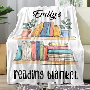 K&ouml;nnte beinhalten: Wei&szlig;e Fleece-Decke mit einem B&uuml;cherregal-Design, das einen Stapel bunter B&uuml;cher und den Text "Emily's reading blanket" zeigt.