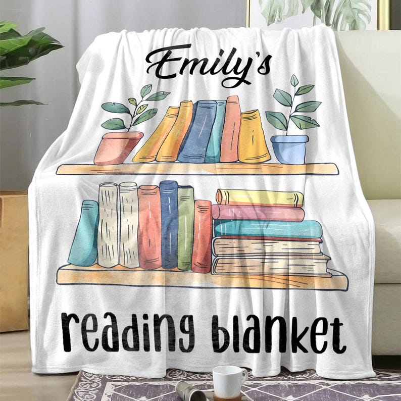 K&ouml;nnte beinhalten: Wei&szlig;e Fleece-Decke mit einem B&uuml;cherregal-Design, das einen Stapel bunter B&uuml;cher und den Text "Emily's reading blanket" zeigt.