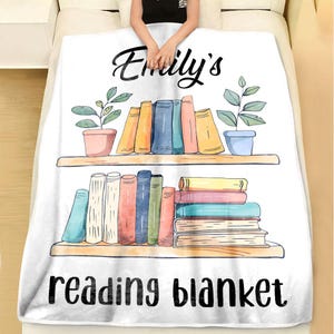 K&ouml;nnte beinhalten: Wei&szlig;e Fleece-Decke mit dem Text "Emily's reading blanket" in Schwarz gedruckt. Die Decke zeigt eine farbenfrohe Illustration von zwei B&uuml;cherregalen mit B&uuml;cherstapeln und Topfpflanzen.