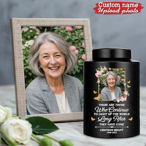 May include: A black memorial urn with a photo of a woman, surrounded by pink flowers and butterflies. Text reads "There are those who continue to light up the world long after they have gone." A framed photo of the same woman is next to the urn.