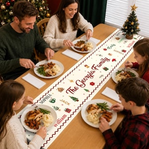 May include: A white fabric table runner featuring a repeating pattern of Christmas-themed icons including trees, gifts, and gingerbread houses, with the text The Georgia Family and various names printed in red, green, and gold script and block fonts.