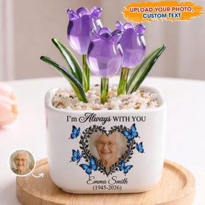 Op de afbeelding: Een witte keramische pot met een foto, een hartvormige krans en blauwe vlinders. De pot bevat drie paarse glazen tulpenbloemen. Tekst op de pot luidt "I'm Always WITH YOU" en "Emma Smith (1945-2026)".
