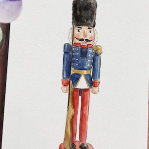 May include: Watercolor painting of a traditional nutcracker soldier. The soldier wears a blue jacket with gold accents, red pants, and a tall black hat. He holds a rifle and stands on a red base. The artwork is on white paper.