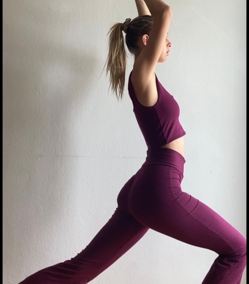 May include: A woman wearing a burgundy sleeveless crop top and matching flared pants. She is standing in a yoga pose with her right leg extended forward.