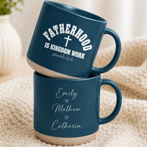 Puede incluir: Dos tazas de cerámica azul marino apiladas. La taza superior tiene el texto "FATHERHOOD IS KINGDOM WORK" con una cruz y "proverbios 22:6". La taza inferior tiene los nombres "Emily", "Mathew" y "Catherin" con corazones.