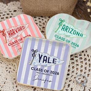 Puede incluir: Tres platos decorativos de cer&aacute;mica con borde dorado. Uno rosa con texto "HARVARD", otro verde con "ARIZONA" y un tercero morado con "YALE". Cada plato tiene rayas verticales y un dise&ntilde;o de bander&iacute;n con el texto "CLASS OF 2026".