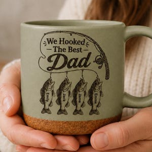 May include: A sage green ceramic mug with a speckled cork base. The front features a dark brown graphic of a fishing rod and four fish, each labeled with the names Emily, Jack, Jame, and Emme, and the text We Hooked The Best Dad.
