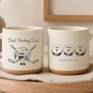 Pode incluir: Duas canecas de cerâmica em tom creme com base de argila natural. A caneca da esquerda apresenta um gráfico preto de um capacete de hóquei e tacos cruzados com o texto Best Hockey Dad. A da direita mostra três discos de hóquei com nomes.
