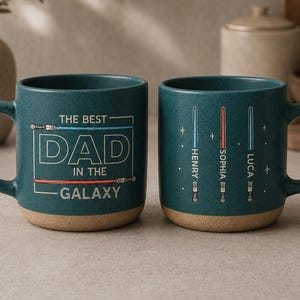 May include: Two teal ceramic mugs with light brown bases. One mug reads "THE BEST DAD IN THE GALAXY" in white text with red and blue lines. The other mug has three lightsaber designs with names. A great gift.
