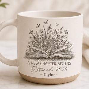 May include: Cream-colored ceramic mug with a brown base and handle. The mug features a black and white illustration of an open book with flowers, butterflies, and the text "A NEW CHAPTER BEGINS Retired 2026 - Taylor -."