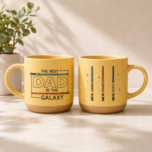 May include: Two yellow ceramic mugs with brown bottoms. One mug reads "THE BEST DAD IN THE GALAXY" with a lightsaber design. The other mug has three lightsaber designs with the names "HENRY", "SOPHIA", and "LUCA".