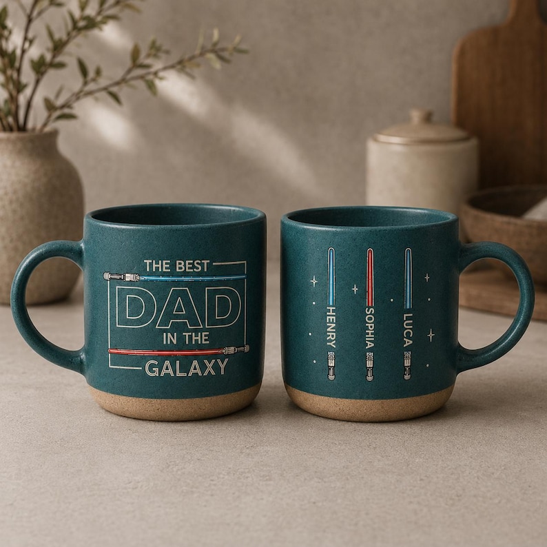 May include: Two teal ceramic mugs with light brown bases. One mug reads "THE BEST DAD IN THE GALAXY" in white text with red and blue lines. The other mug has three lightsaber designs with names. A great gift.