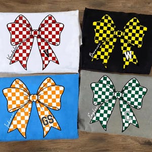 Puede incluir: Cuatro camisetas dobladas con diseños de lazo. Los lazos son de cuadros rojos y blancos, amarillos y negros, naranjas y blancos, y verdes y blancos. Cada lazo tiene una letra y un nombre de equipo.
