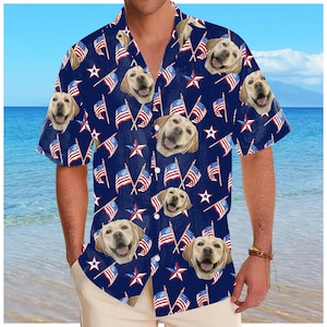 May include: A short-sleeved button-down shirt with a navy blue background. The shirt features a repeating pattern of American flags, red and white stars, and smiling Labrador Retriever faces. The shirt is worn with khaki pants.
