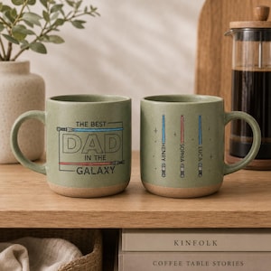 May include: Two green ceramic mugs with a light brown base. One mug has the text "THE BEST DAD IN THE GALAXY" with lightsaber illustrations. The other mug has lightsaber illustrations with names. The mugs are on a wooden shelf.