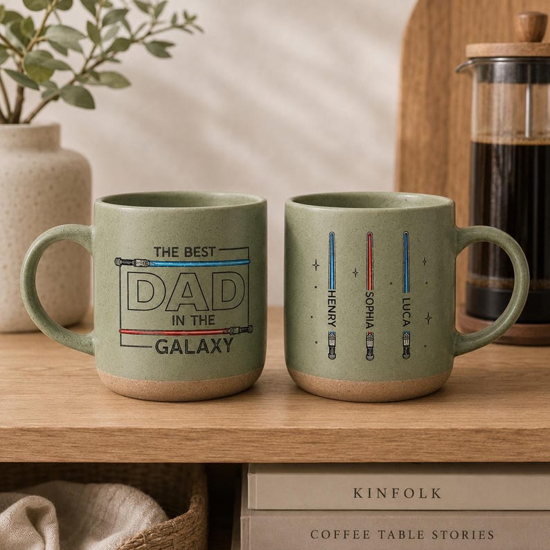 May include: Two green ceramic mugs with a light brown base. One mug has the text "THE BEST DAD IN THE GALAXY" with lightsaber illustrations. The other mug has lightsaber illustrations with names. The mugs are on a wooden shelf.