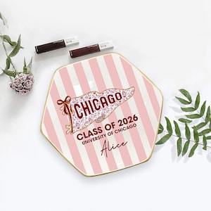 Puede incluir: Un plato decorativo con un patr&oacute;n de rayas rosas y blancas y un borde dorado. El plato presenta un dise&ntilde;o de bander&iacute;n con la palabra "CHICAGO" y el texto "CLASS OF 2026 UNIVERSITY OF CHICAGO Alice". Dos tubos de Maybelline SuperStay Matte Ink est&aacute;n en la parte superior.