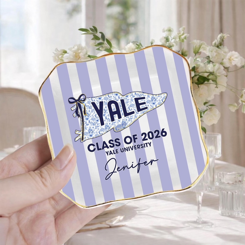Puede incluir: Un plato decorativo con borde dorado y fondo a rayas blancas y moradas claras. El plato presenta el texto "YALE CLASS OF 2026 YALE UNIVERSITY" y un nombre en letra cursiva. El logotipo de Yale est&aacute; en un dise&ntilde;o floral azul.
