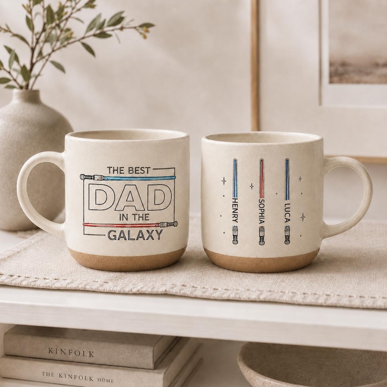 May include: Two cream-colored ceramic mugs with brown bases. One mug reads "THE BEST DAD IN THE GALAXY" with a lightsaber design. The other mug has three lightsaber designs with names: Henry, Sophia, and Luca.