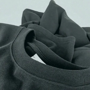 May include: Close-up of a dark gray t-shirt, with a focus on the neckline and collar. The fabric appears soft and slightly textured. Additional folded fabric is visible in the background, suggesting a stack of shirts.
