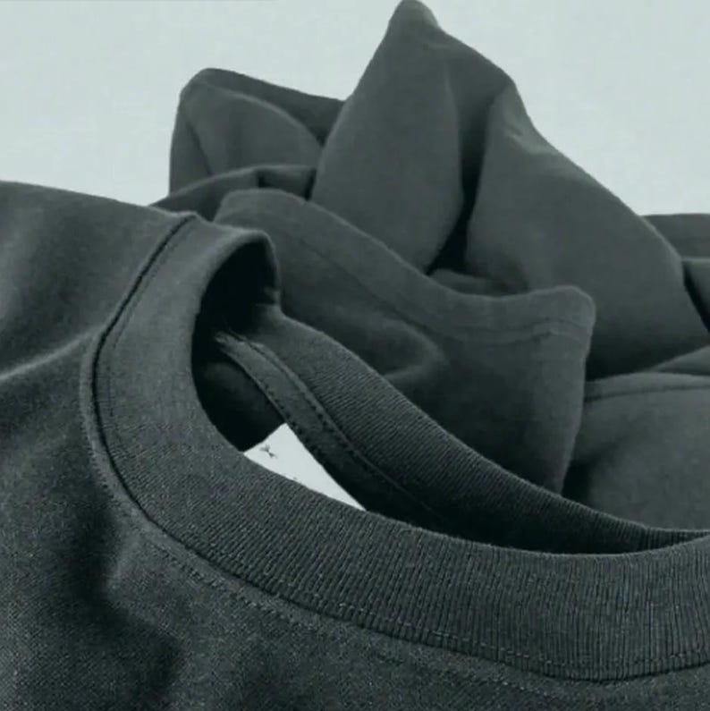 May include: Close-up of a dark gray t-shirt, with a focus on the neckline and collar. The fabric appears soft and slightly textured. Additional folded fabric is visible in the background, suggesting a stack of shirts.