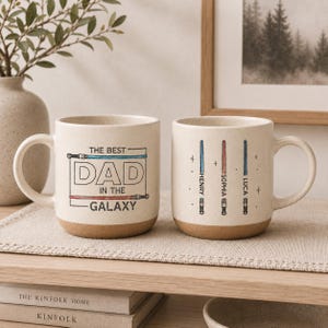 May include: Two beige ceramic mugs with brown bases. One mug reads "THE BEST DAD IN THE GALAXY" in black and blue text. The other mug has three lightsaber designs with names: Henry, Sophia, and Luca.