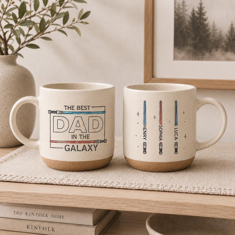May include: Two beige ceramic mugs with brown bases. One mug reads "THE BEST DAD IN THE GALAXY" in black and blue text. The other mug has three lightsaber designs with names: Henry, Sophia, and Luca.
