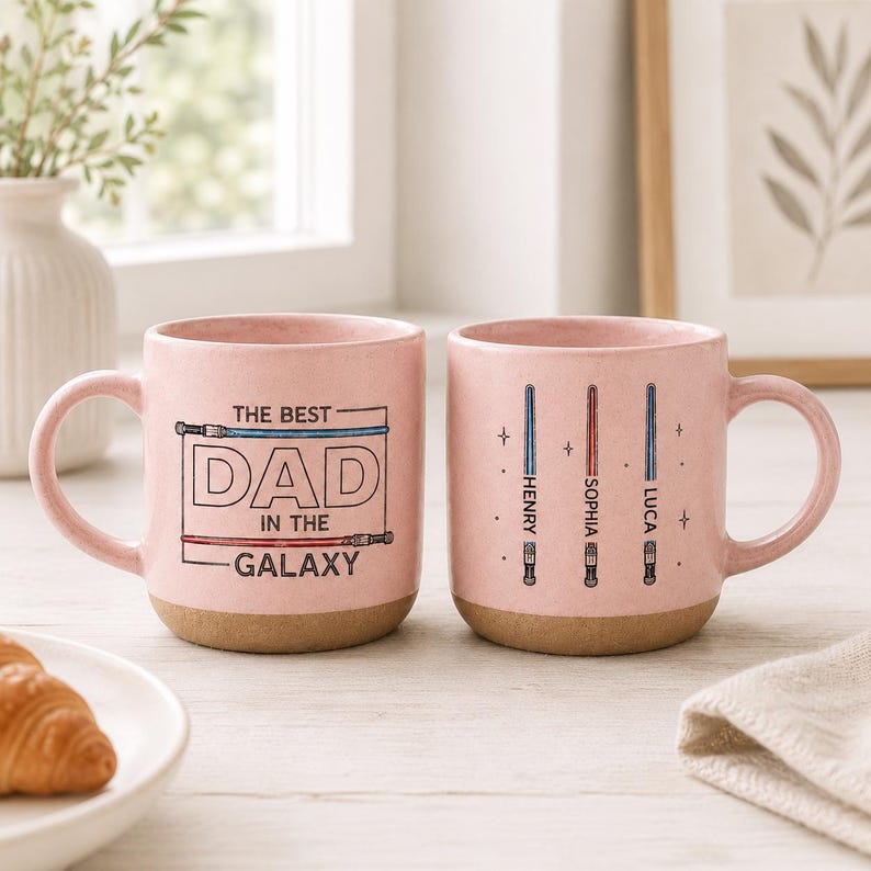 May include: Two pink ceramic mugs with brown bases. One mug reads "THE BEST DAD IN THE GALAXY" in black text with a blue and red lightsaber design. The other mug has three lightsaber designs with the names "HENRY", "SOPHIA", and "LUCA".