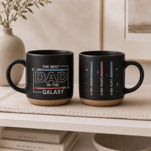 May include: Two black ceramic mugs with a light brown base. One mug has the text "THE BEST DAD IN THE GALAXY" in white. The other mug has three lightsaber designs with names. Both mugs have a curved handle.