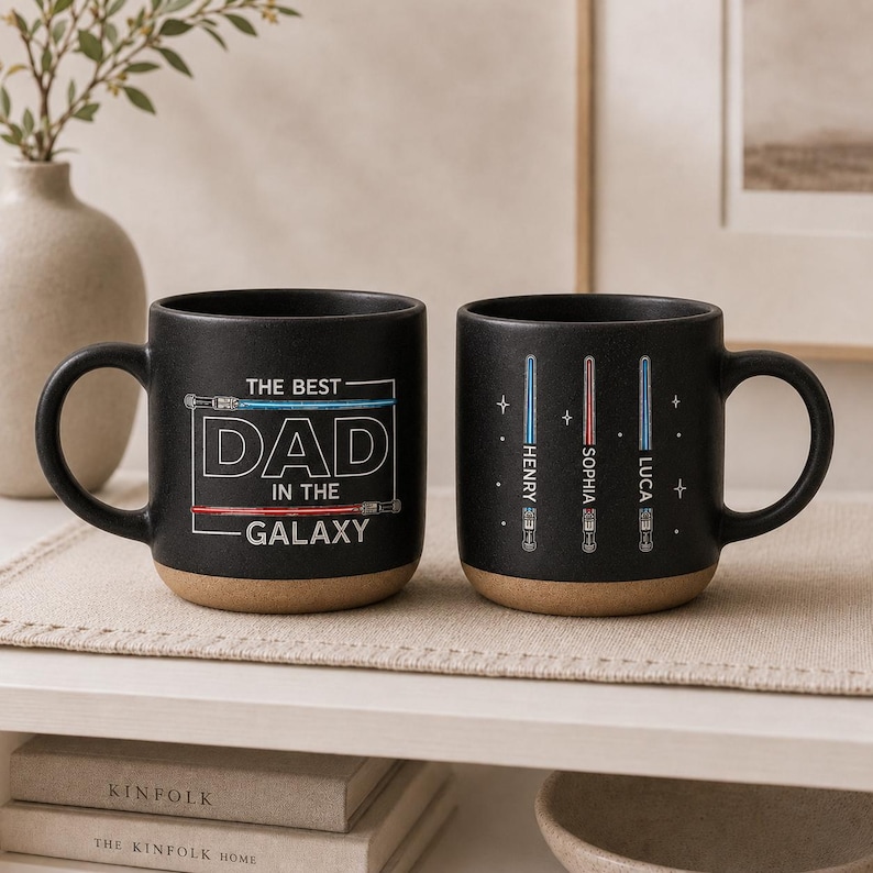 May include: Two black ceramic mugs with a light brown base. One mug has the text "THE BEST DAD IN THE GALAXY" in white. The other mug has three lightsaber designs with names. Both mugs have a curved handle.