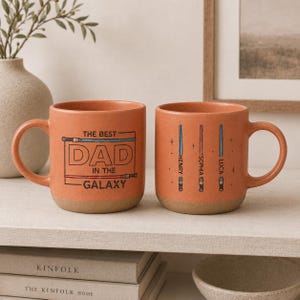 May include: Two orange ceramic mugs with light brown bases. One mug has the text "THE BEST DAD IN THE GALAXY" in a black outline. The other mug has three lightsaber designs with names. A great gift for a dad.
