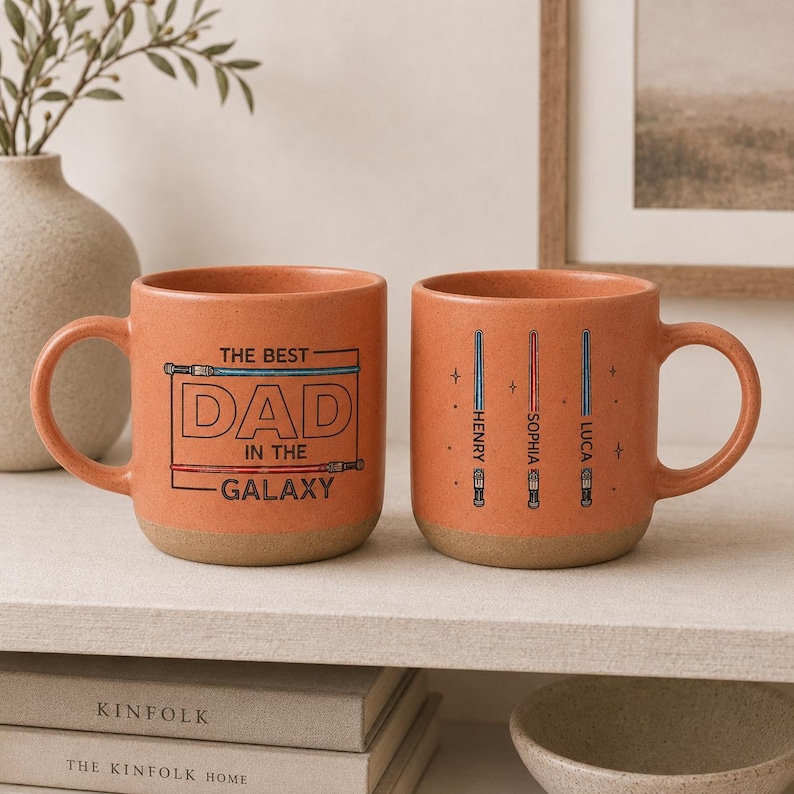 May include: Two orange ceramic mugs with light brown bases. One mug has the text "THE BEST DAD IN THE GALAXY" in a black outline. The other mug has three lightsaber designs with names. A great gift for a dad.
